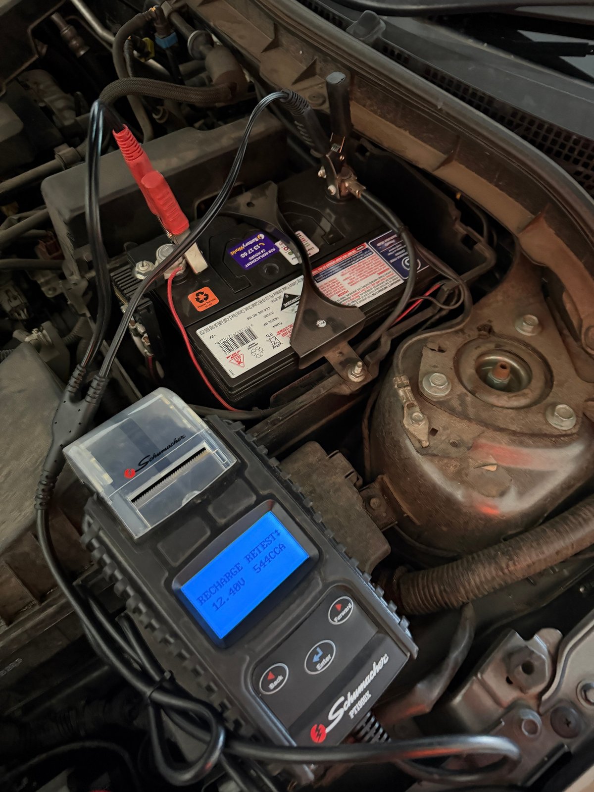 Car battery with jumper cables connected and diagnostic scanner with blue screen resting on engine compartment